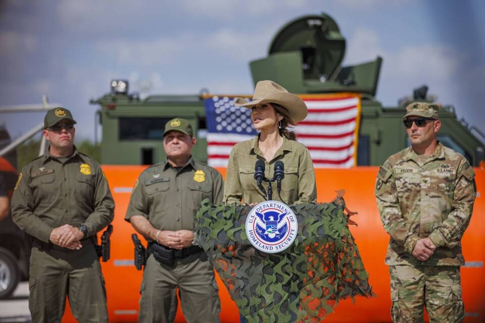 A woman in a cowboy hat stands at a podium with men in military-style garb and military equipment behind her. 