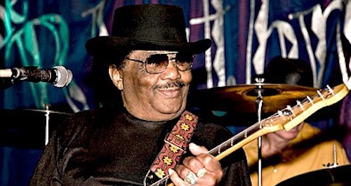 Seated man wearing black hat and playing electric guitar