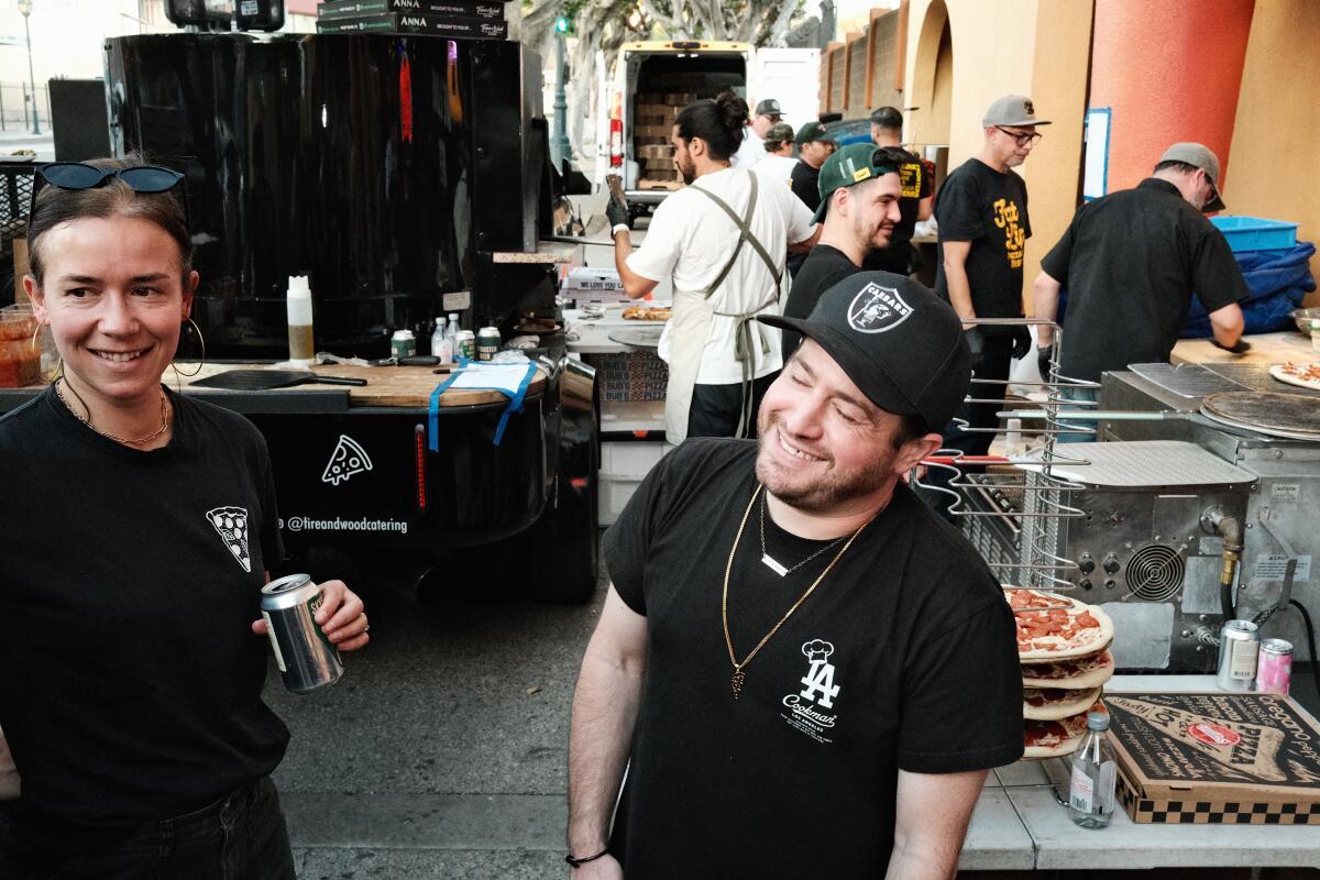 A woman and a man stand outside a busy restaurant where multiple outdoor pizza ovens are set up