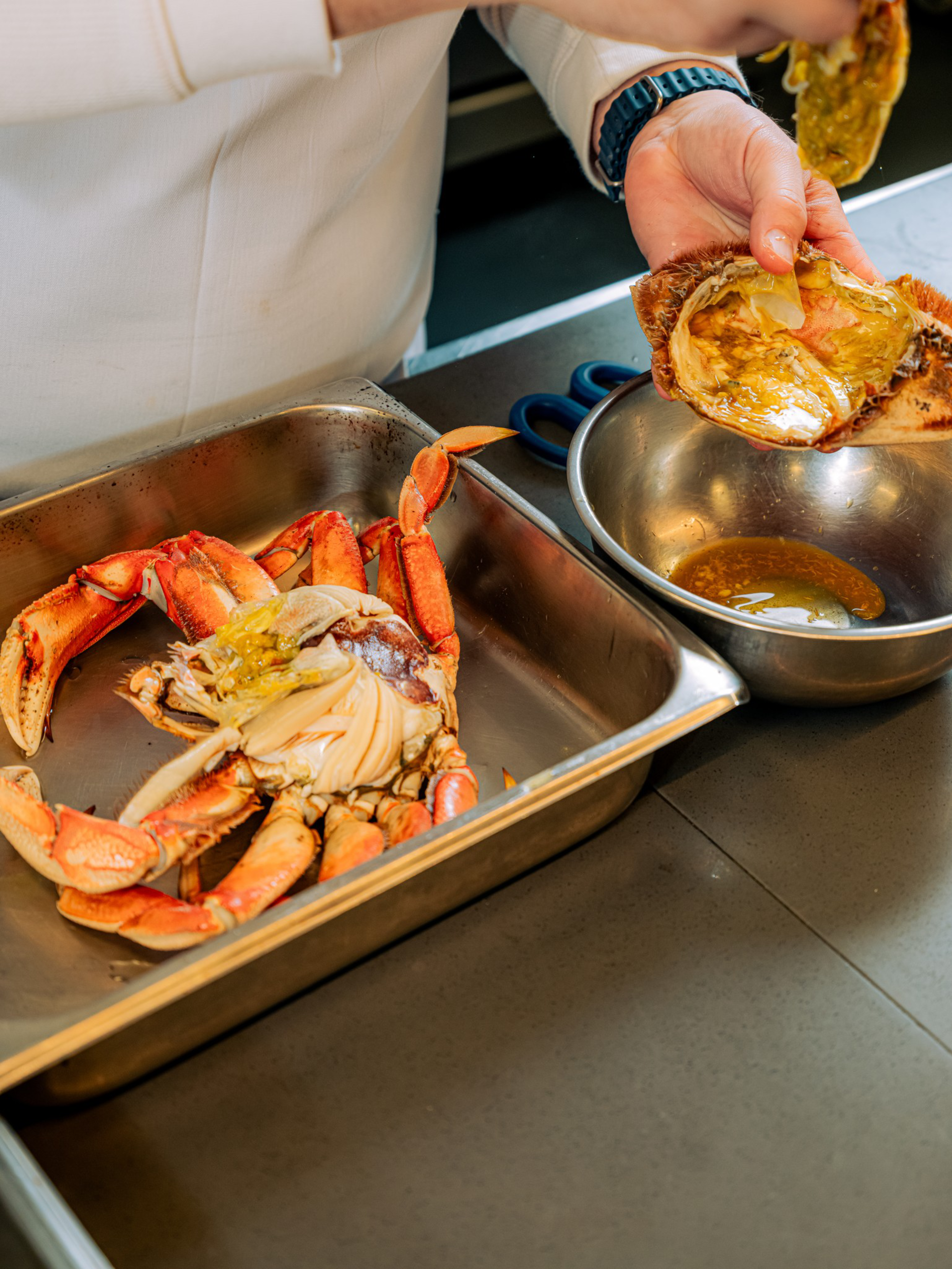 A person is cleaning and preparing a cooked crab, removing its shell and placing the contents into a metal bowl beside a tray holding the crab’s body.