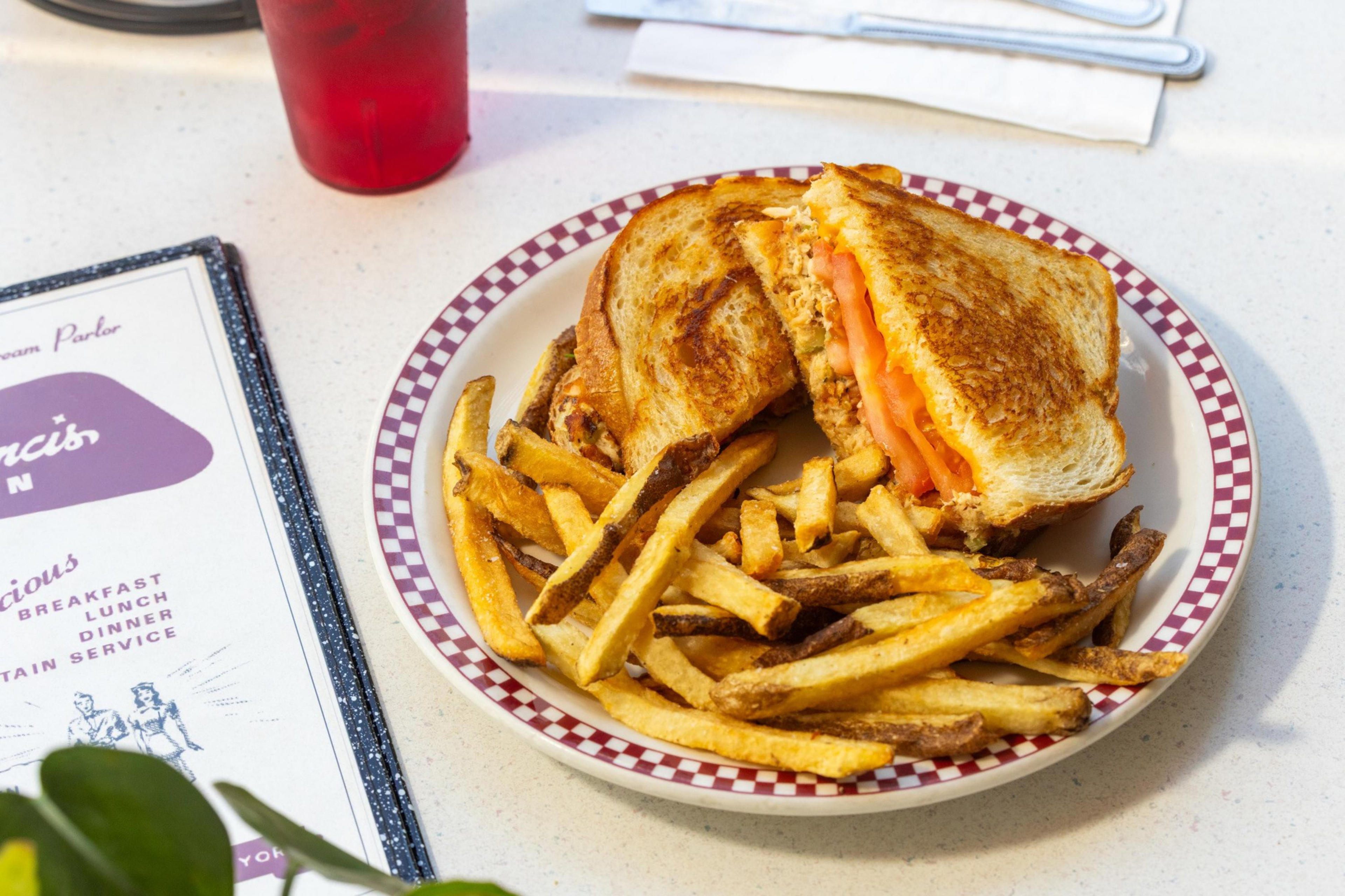 A grilled cheese sandwich with tomato slices is served with crispy French fries on a checkered-edge plate, next to a red drink and a menu.