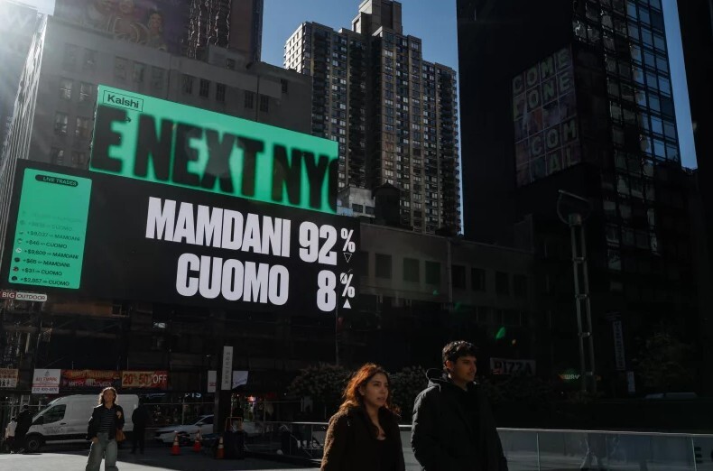 two people walk along a city street while in the background a large digital billboard has the words "Mamdani 92% Cuomo 8%" on it
