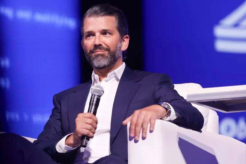 a white man with dark hair in a blue suit sits on a stage and holds a microphone