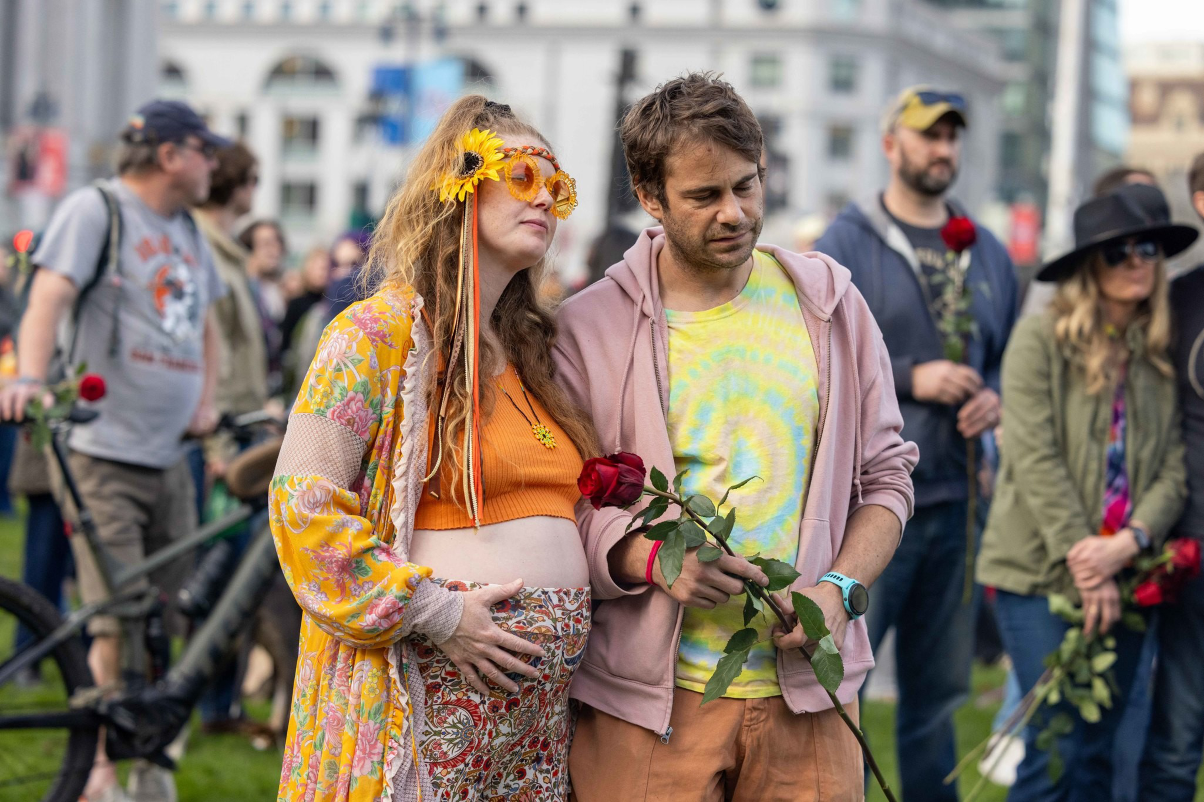 A pregnant woman and a man stand closely, each holding a red rose, surrounded by people in casual attire at an outdoor gathering.
