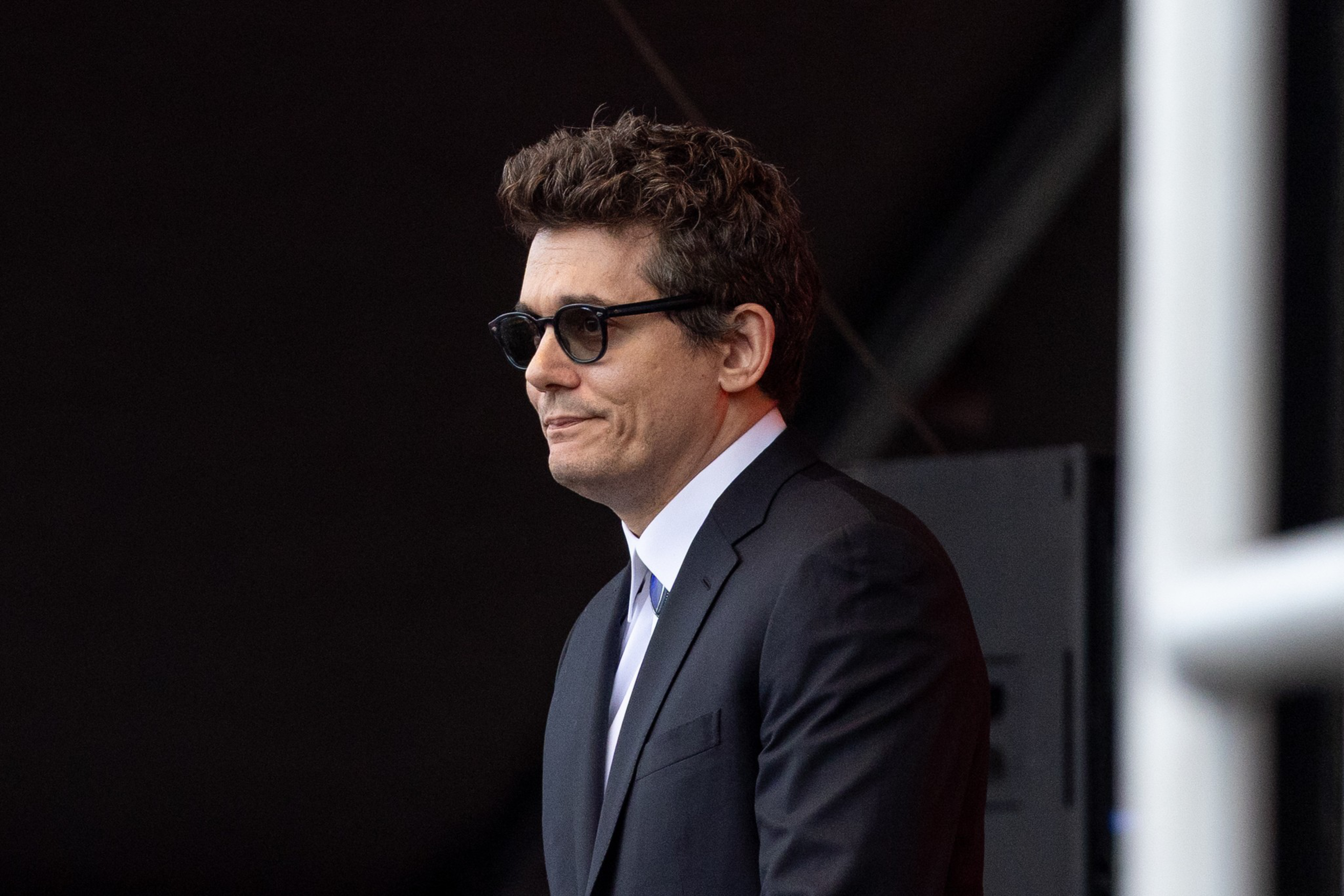 A man with short curly hair and dark sunglasses wears a black suit with a white shirt, standing against a dark blurred background.