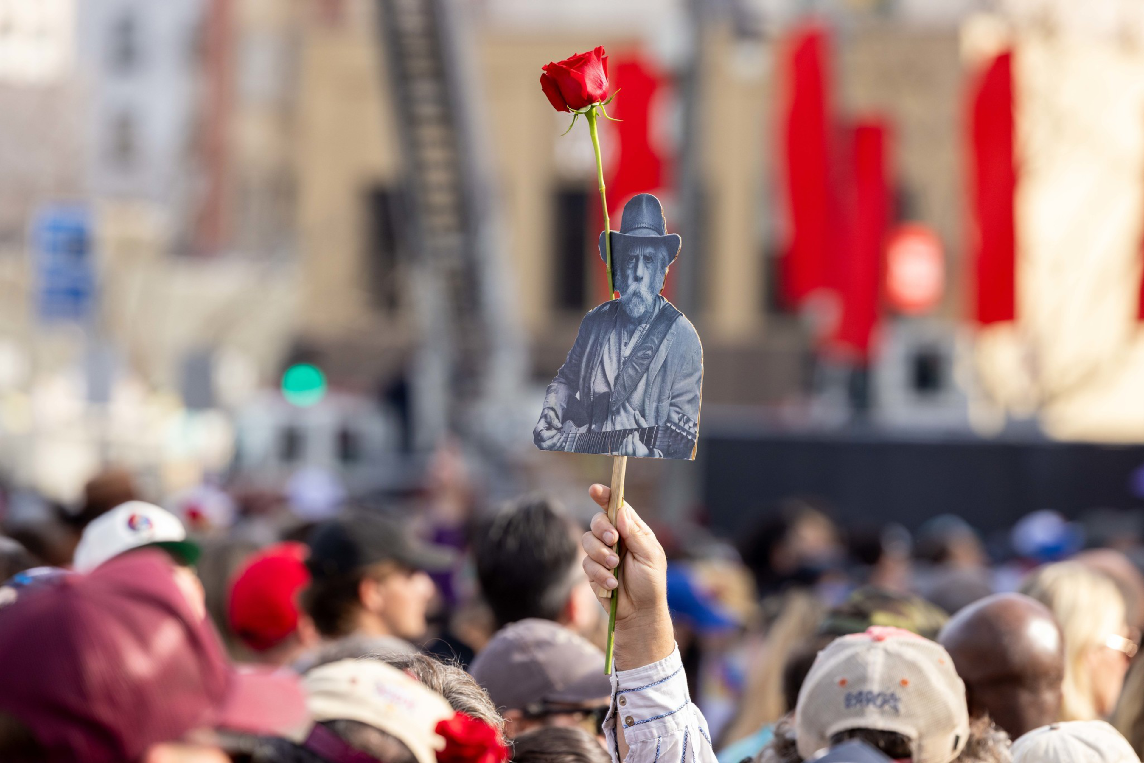 A hand holds a stick with a black-and-white photo of a man wearing a hat and playing guitar, topped with a single red rose, above a crowd.