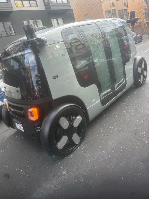 A self-driving electric vehicle with large black wheels and sliding glass doors is parked on a city street.