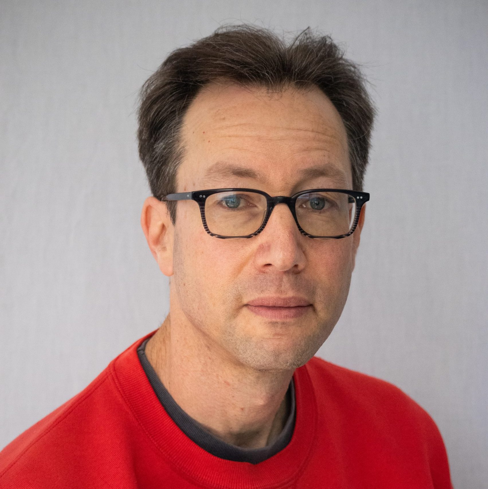A man with short brown hair and glasses wearing a red sweatshirt looks at the camera against a plain light background.