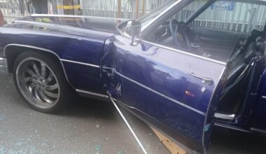 A blue car with a damaged door and shattered glass on the ground; the door is ajar and a metal rod lies beside it. People and barriers are visible in the background.