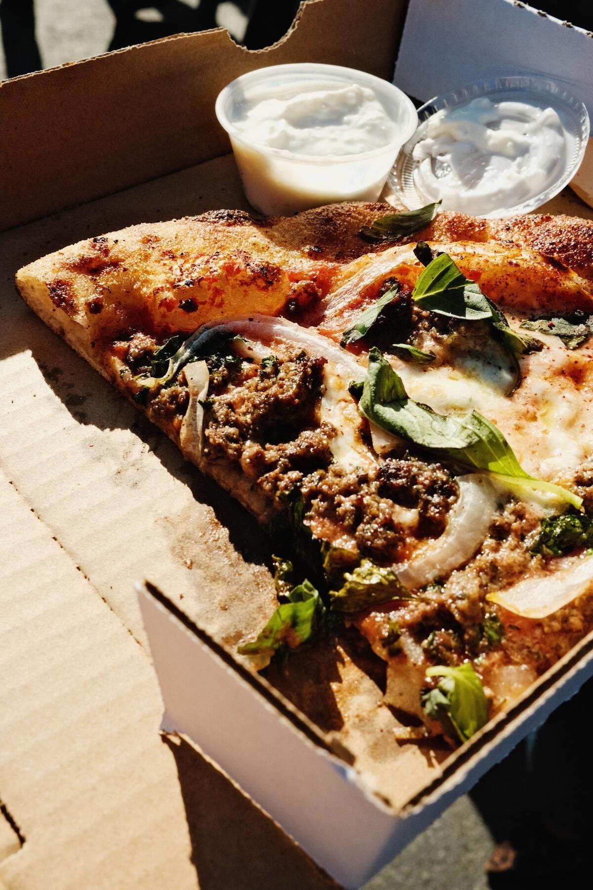 A large slice of koobideh and herb pizza with a side of garlic sauce in a cardboard box