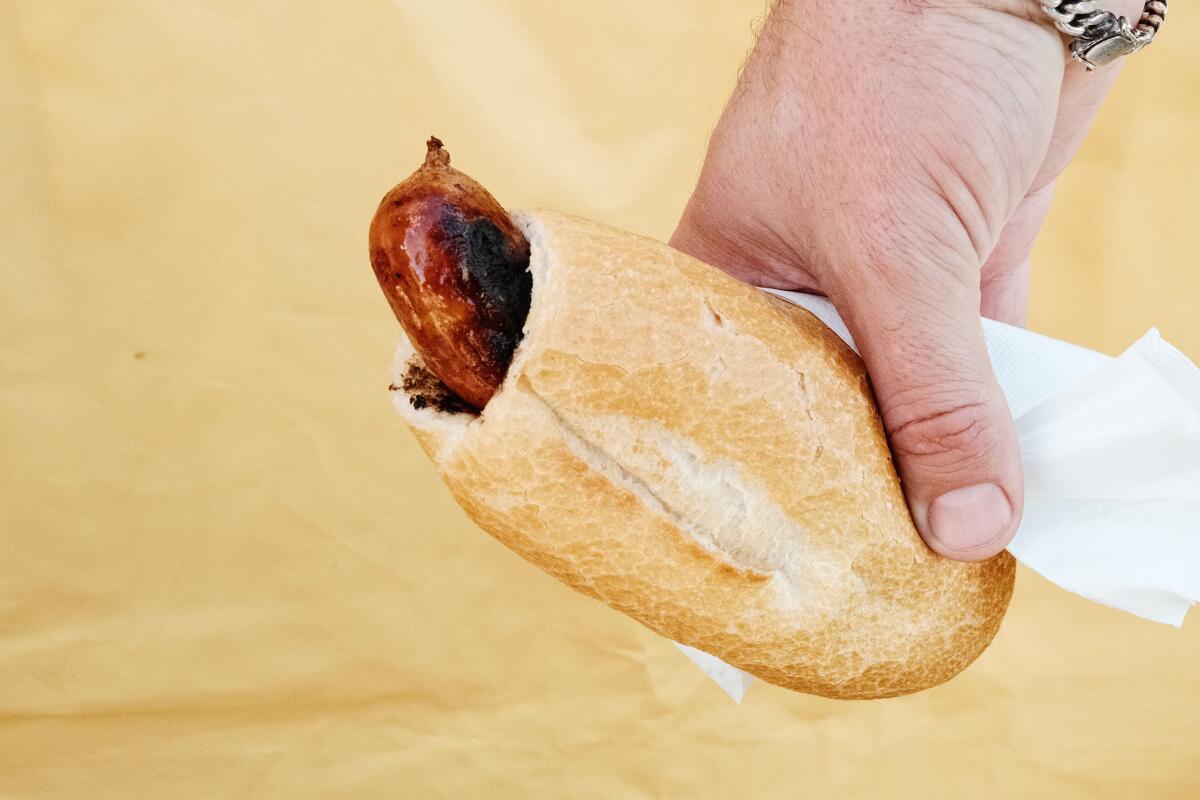 A hand holds a baguette filled with a cheese-stuffed bratwurst against a pale yellow wall