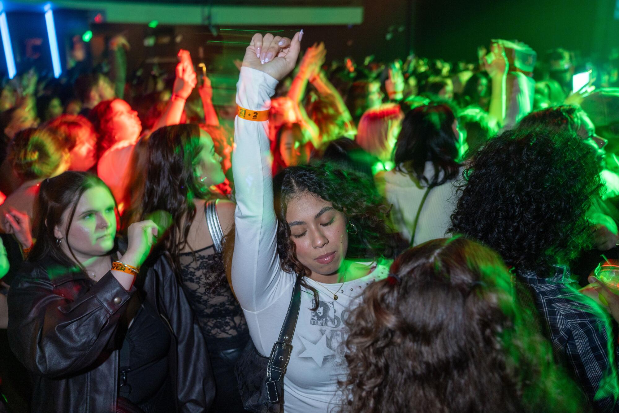 A woman in a white long sleeve top holds an arm up as she's surrounded by a crowd of people dancing.