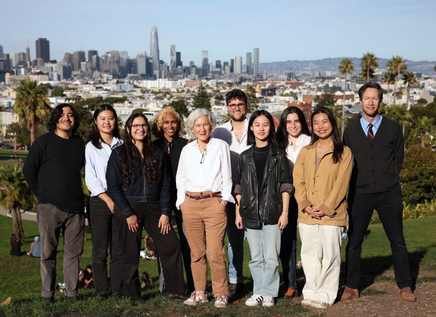 A group of ten people standing outdoors in a park with a city skyline in the background.
