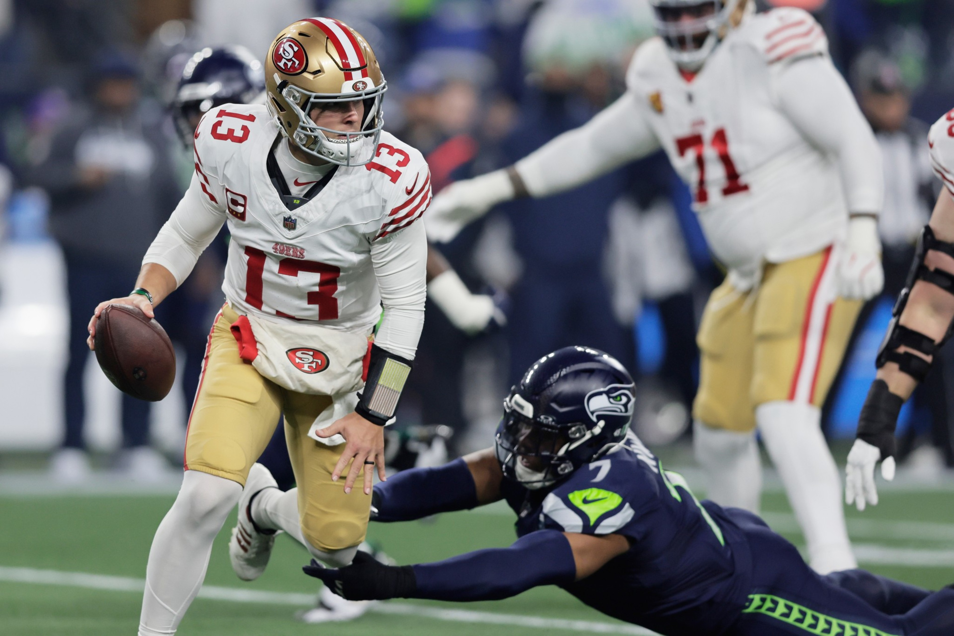 A San Francisco 49ers quarterback with the ball evades a diving Seattle Seahawks defender during a football game.