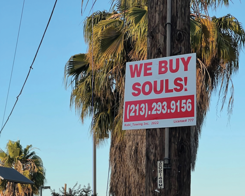 A sign reading "We Buy Souls" on a telephone pole in front of a palm tree. 