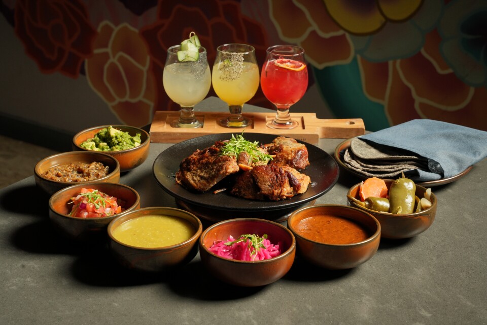 A taco bar setup with sauces, meat, tortillas and three cocktails.