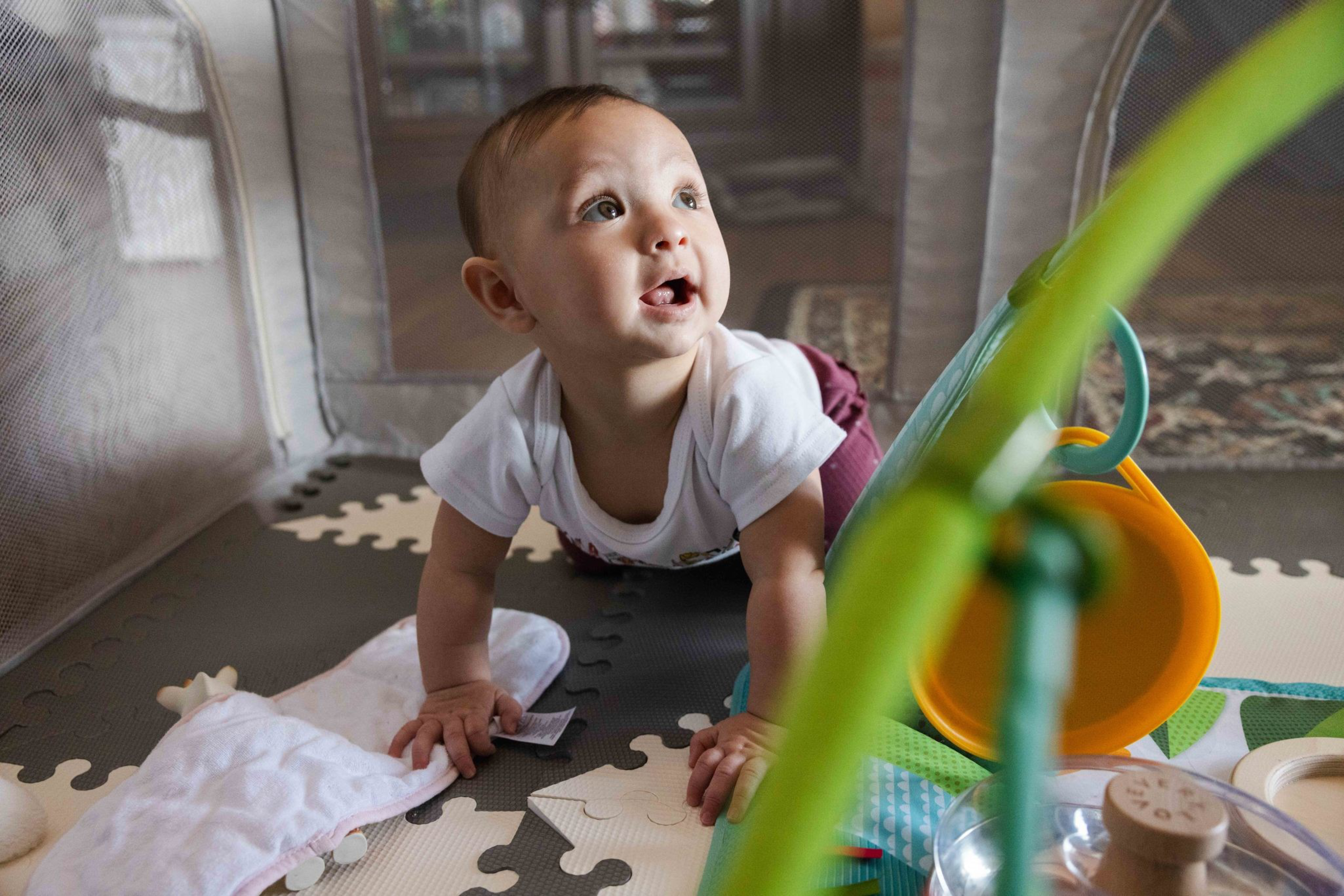 A baby in a white shirt is crawling on a foam play mat, looking up with curiosity, surrounded by colorful toys in a playpen.