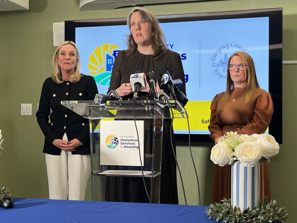Sarah Mahin (center), a woman with light skin tone, speaks at a podium about the launch of the new county homelessness department she will direct. Standing behind her are L.A. County Supervisors Kathryn Barger and Lindsey Horvath, two women with light skin tone.