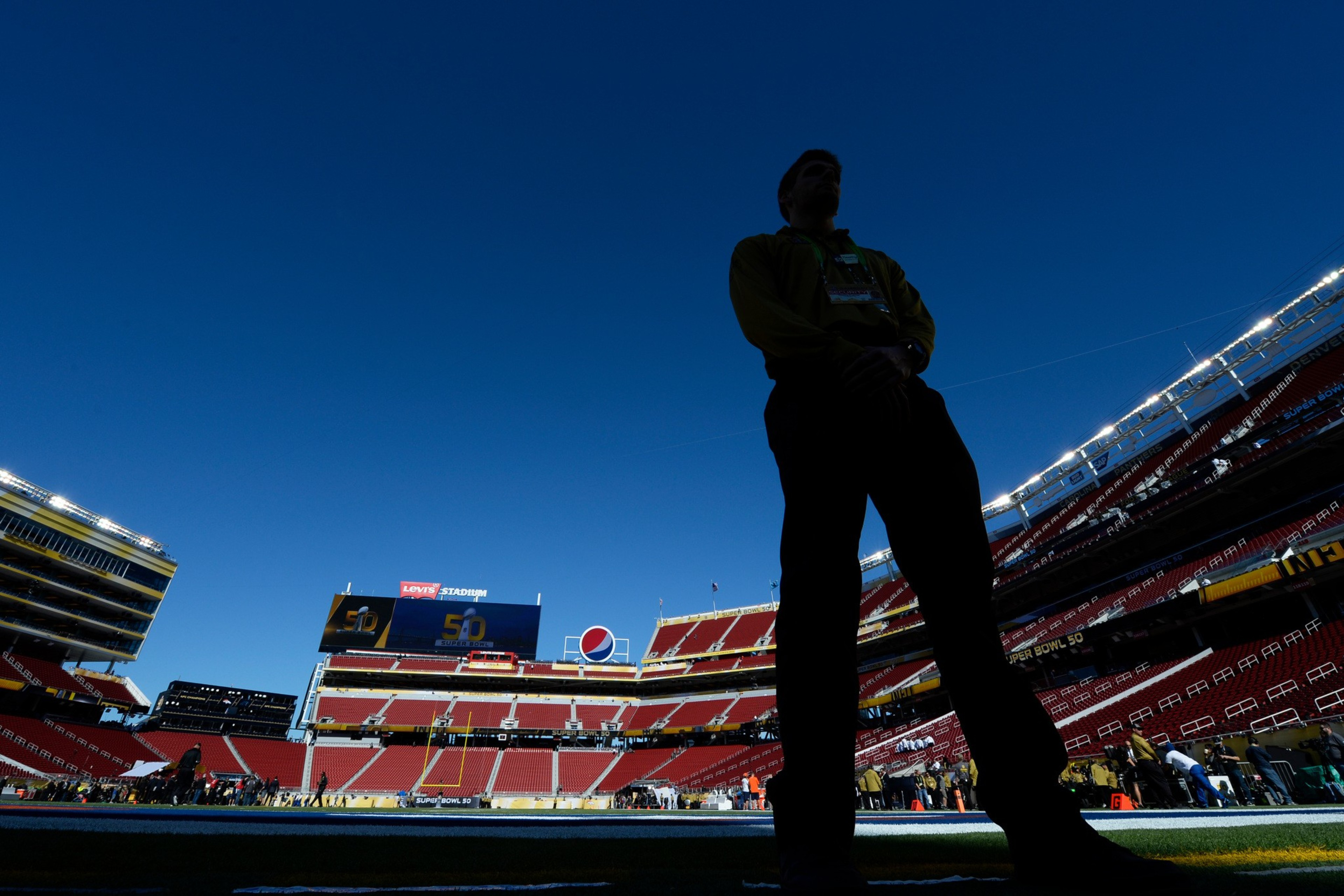 A person stands in silhouette on a stadium field under a clear blue sky, with empty red seats and a scoreboard displaying “Levi’s Stadium” and the number 50.