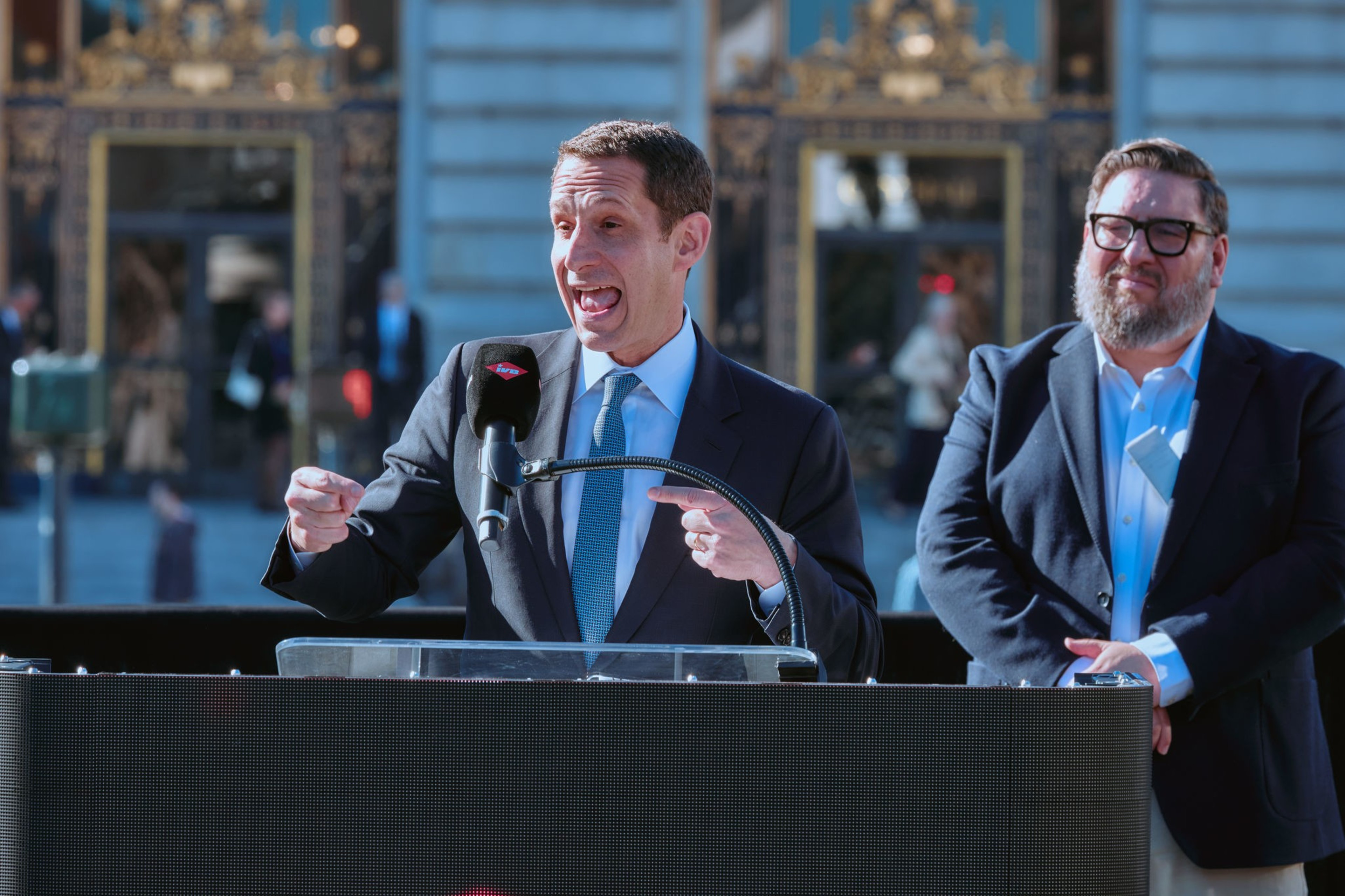 A man in a suit and blue tie speaks animatedly at a microphone, while another man in glasses and a dark jacket stands behind him, smiling.