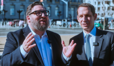 A bearded man in sunglasses and a suit smiles in front of a large domed government building under a clear blue sky.