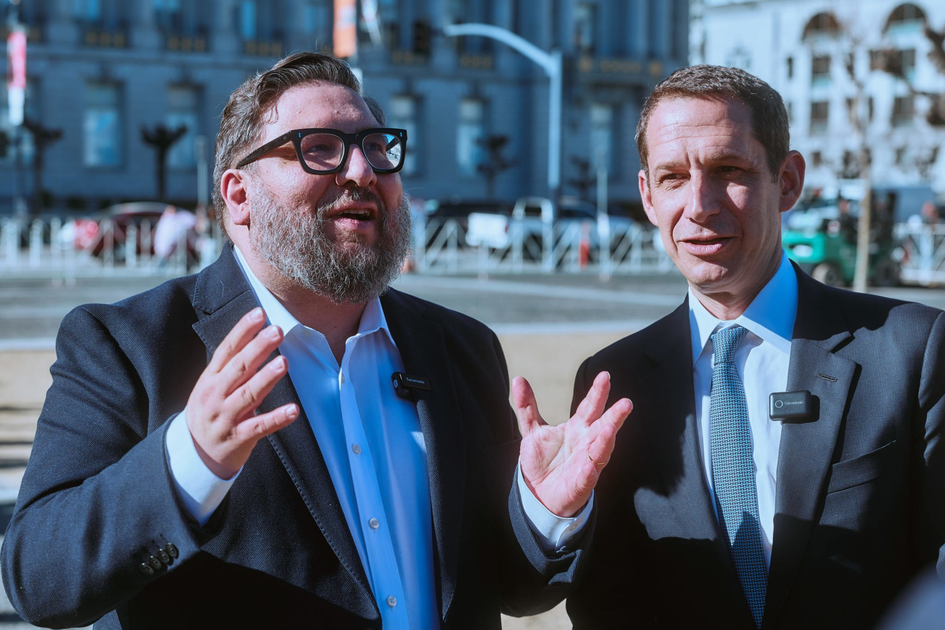 A bearded man in sunglasses and a suit smiles in front of a large domed government building under a clear blue sky.