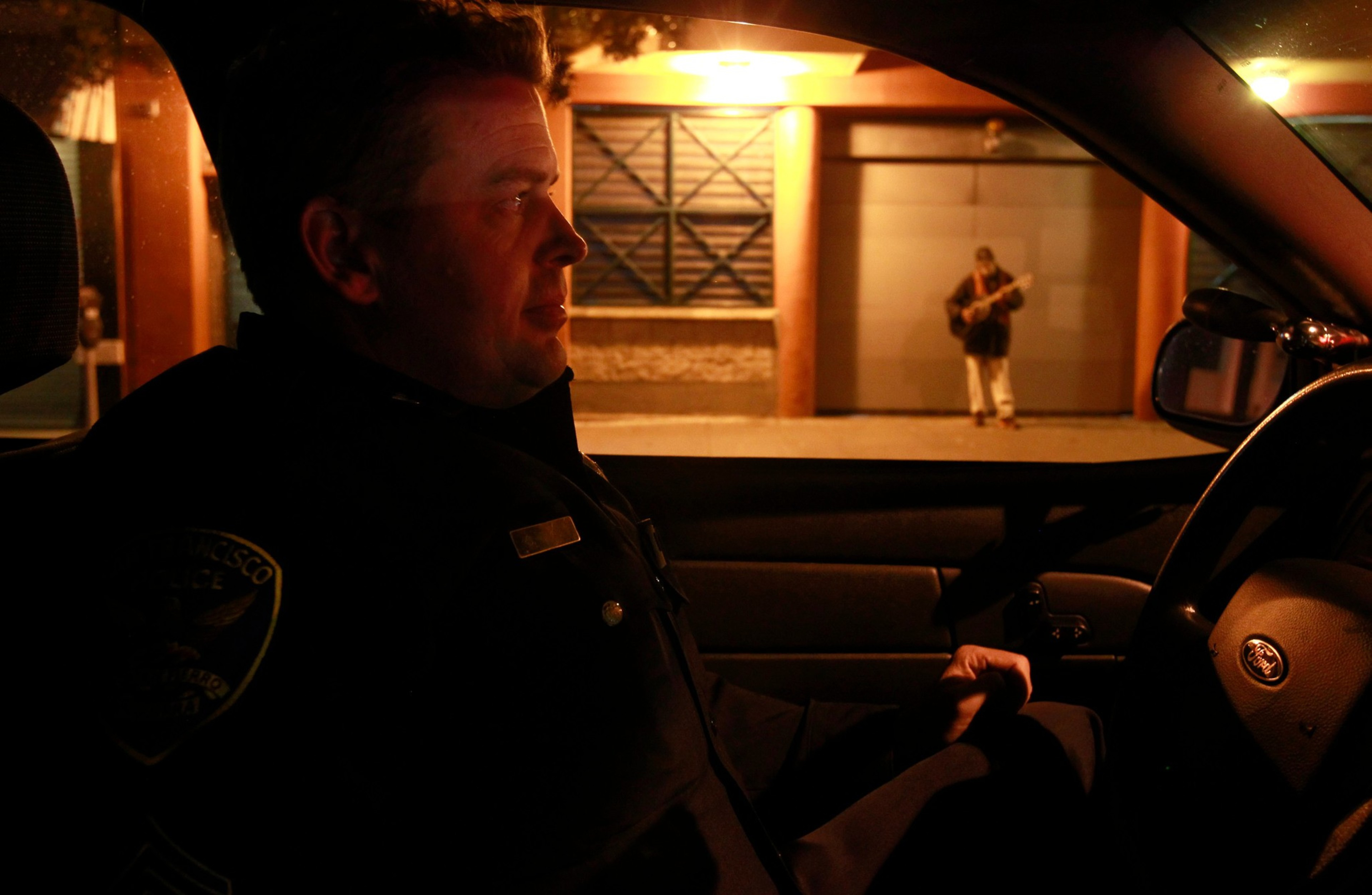 A police officer sits inside a patrol car at night, looking through the window at a person playing guitar on the sidewalk.