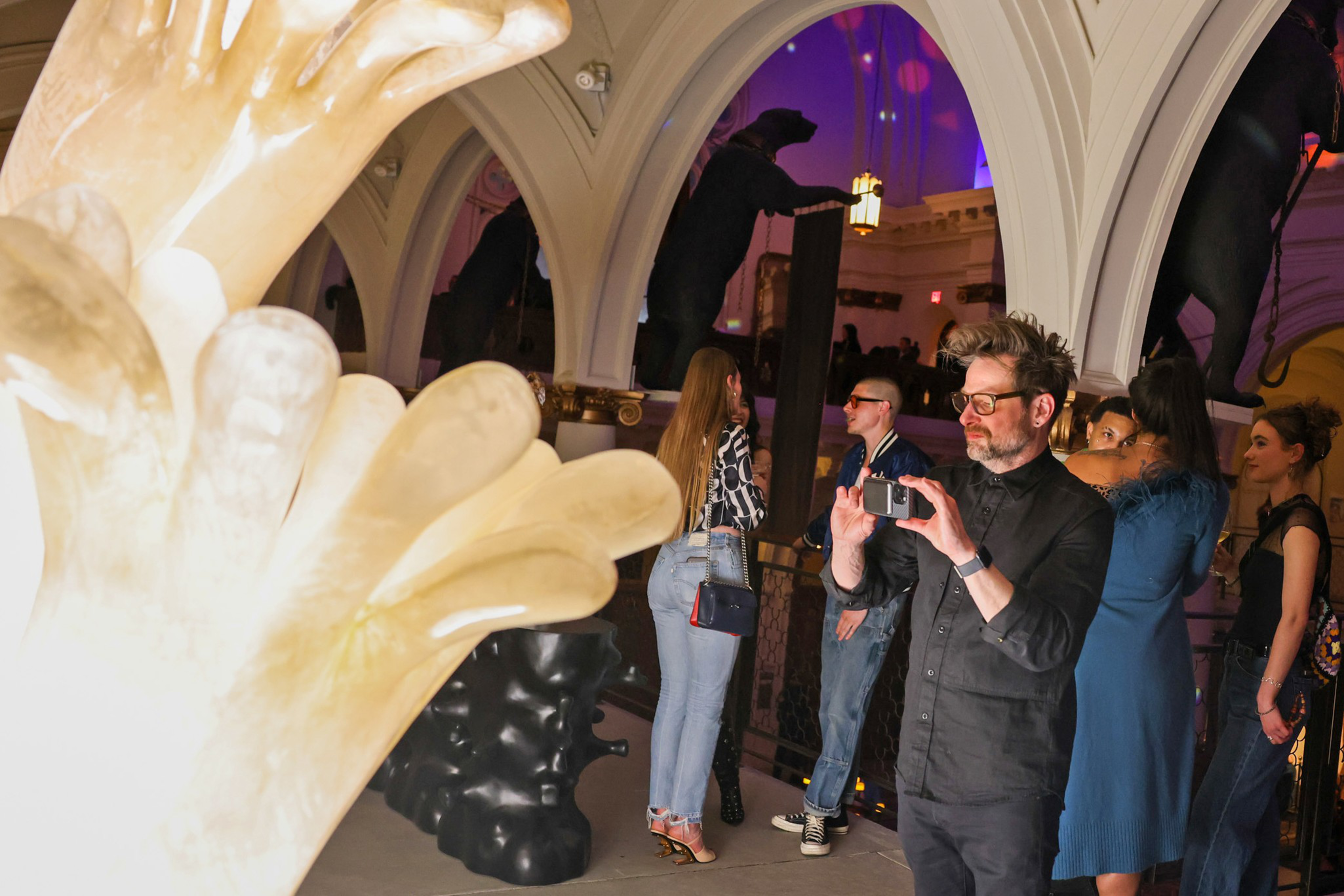 A group of people stand in an arched room with decorative black bear sculptures, while a man in dark clothes and glasses takes a photo.