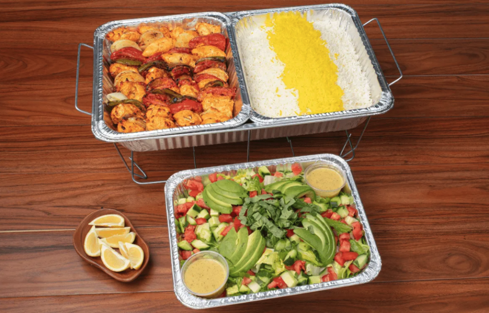 Three foil containers sit on a wooden table. In one is an array of kabobs; in another a mix of yellow and white reice, and in the third a salad of chopped cucumbers and tomatoes topped with slices of avocado