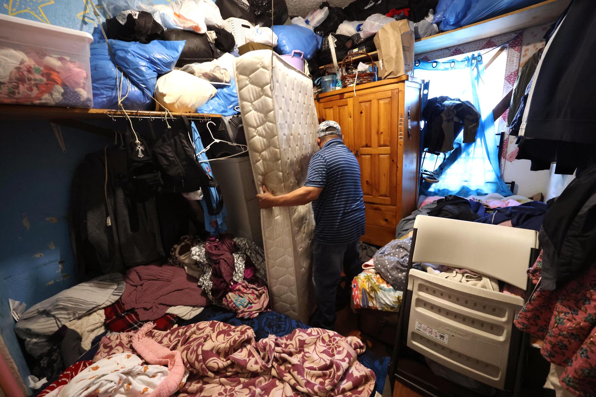 A man carries a mattress in a cluttered apartment.