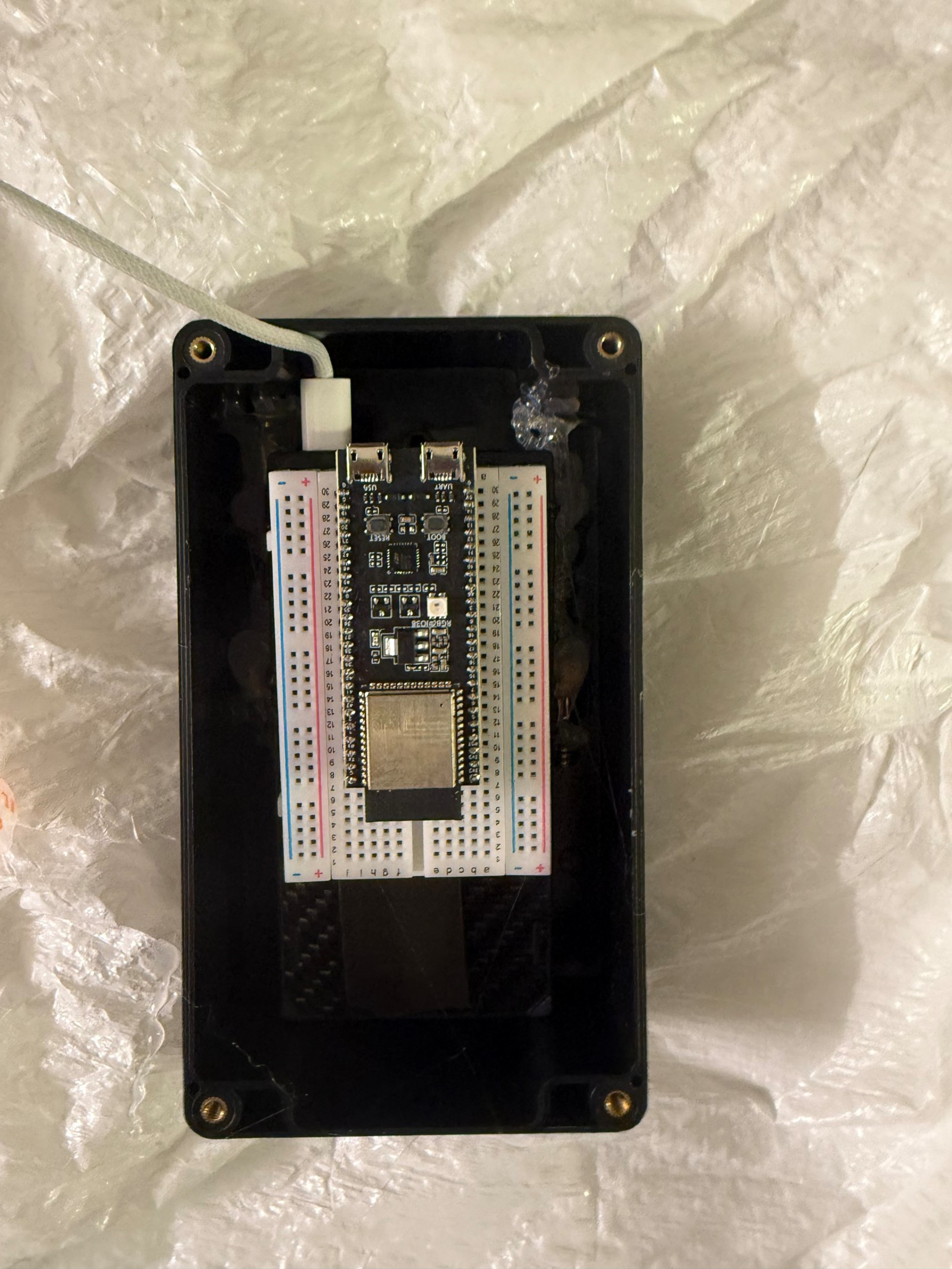 A black rectangular case contains a small white breadboard with an ESP32 microcontroller mounted on it, connected via a white USB cable.