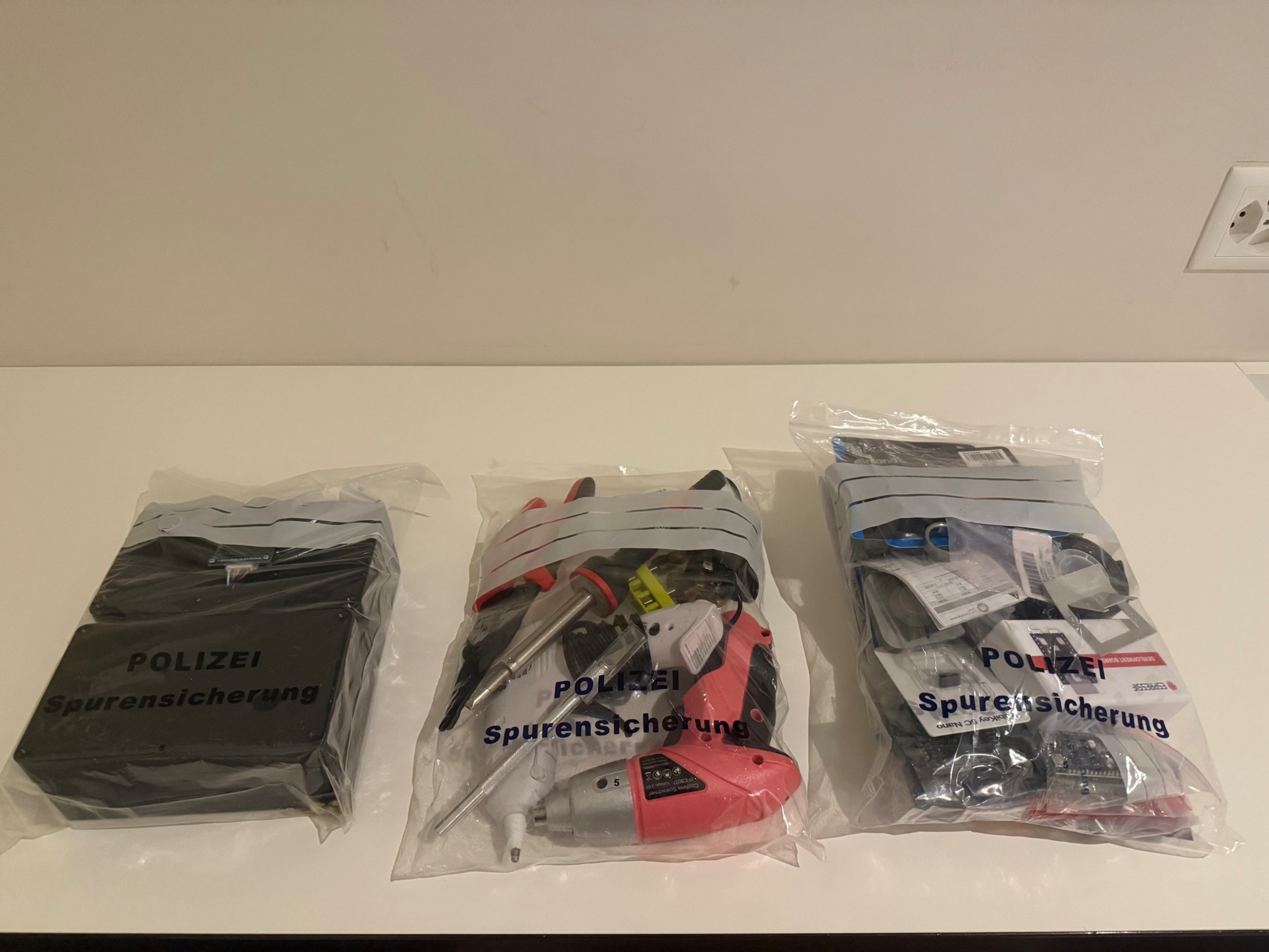 Three clear plastic evidence bags labeled “POLIZEI Spurensicherung” contain various items including a black box, tools like a drill and screwdriver, and miscellaneous small objects.