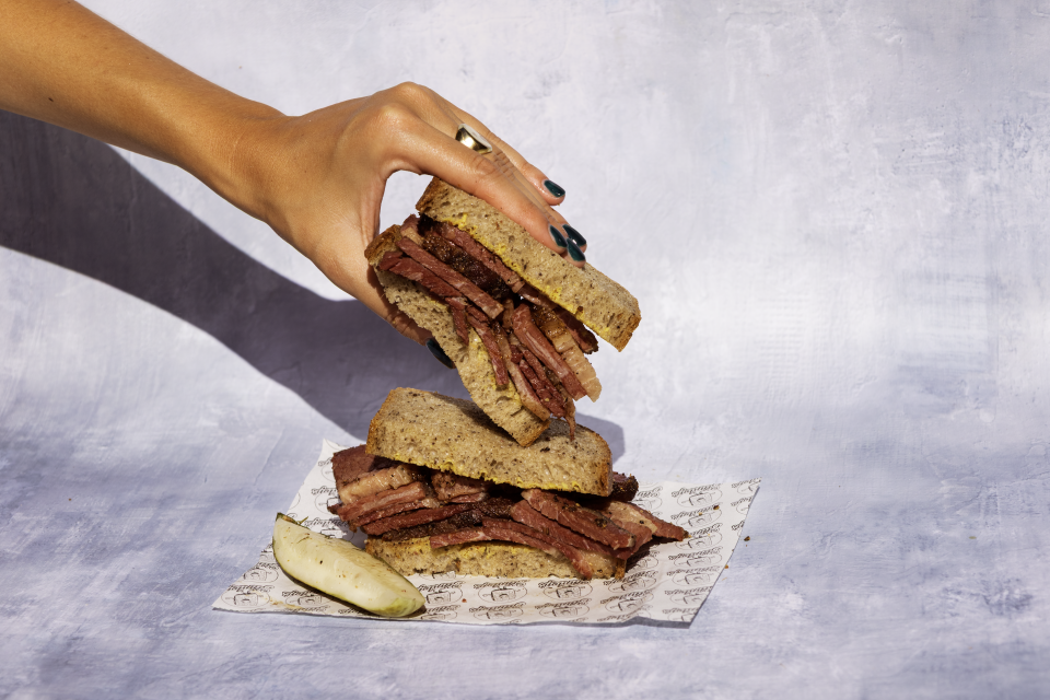 A hand holds the top half of a pastrami sandwich on sourdough bread, revealing thick-cut pink and brown pastrami slices with visible smoke rings and fat marbling, served with a pickle.