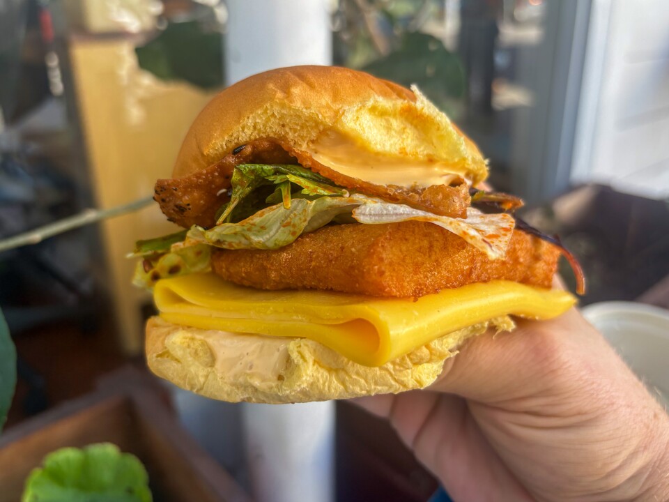 A hand with a light skin tone holds a breakfast sandwich on a potato bun filled with a fried croquette, yellow steamed egg layer, American cheese, and fresh veg.