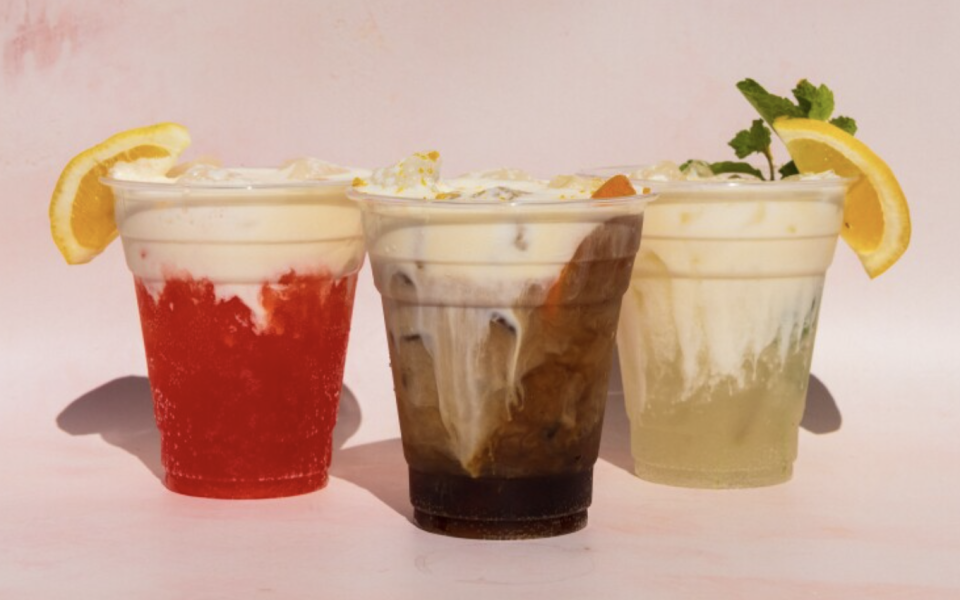 Three plastic cups contain colorful drinks, one red, one brown, one yellow, with a creamy top; each are garnished with ice and an orange slice