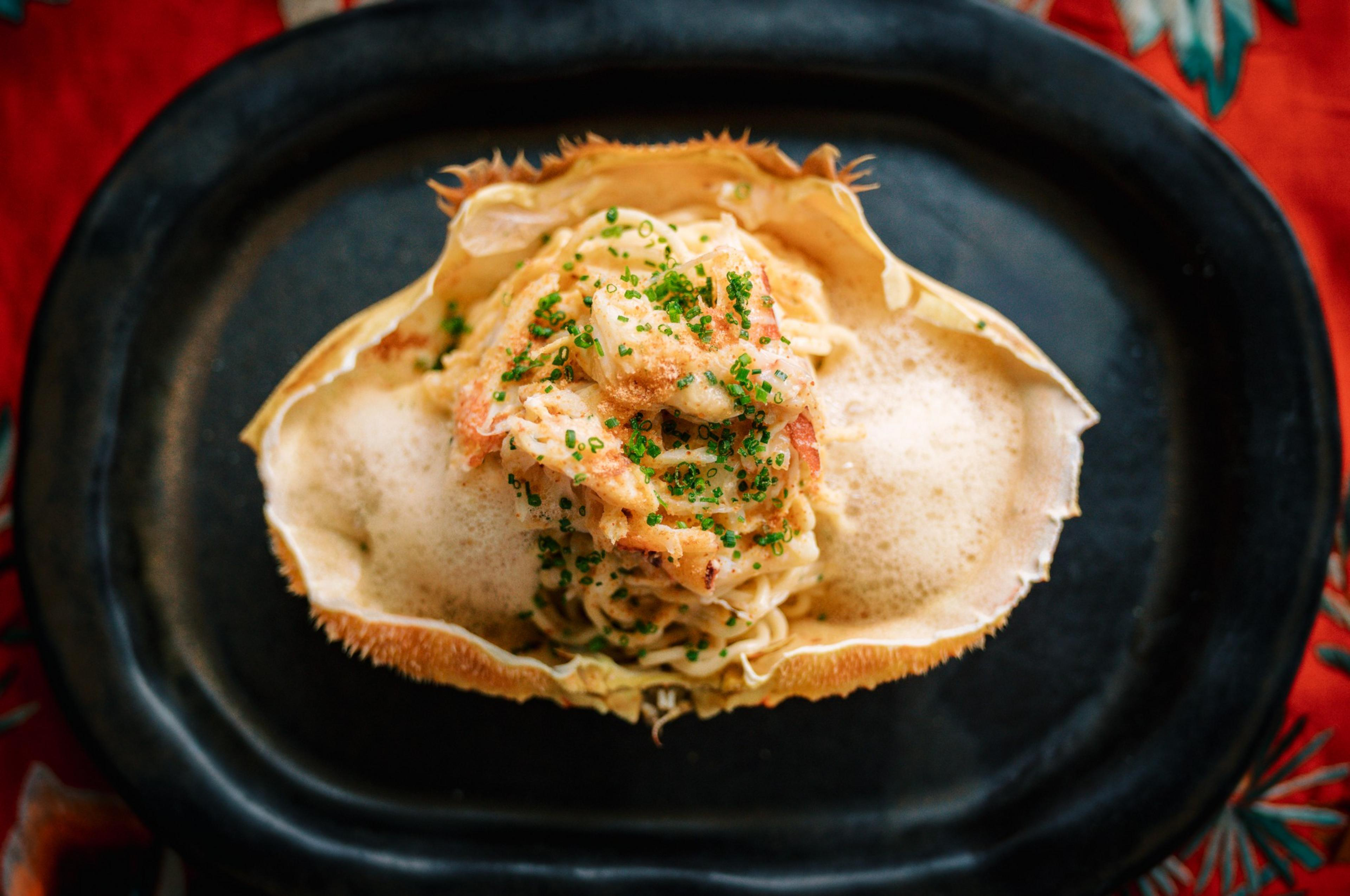 Cooked crab meat noodles garnished with green herbs rest inside a crab shell, served on a black oval plate with a foamy sauce on the sides.