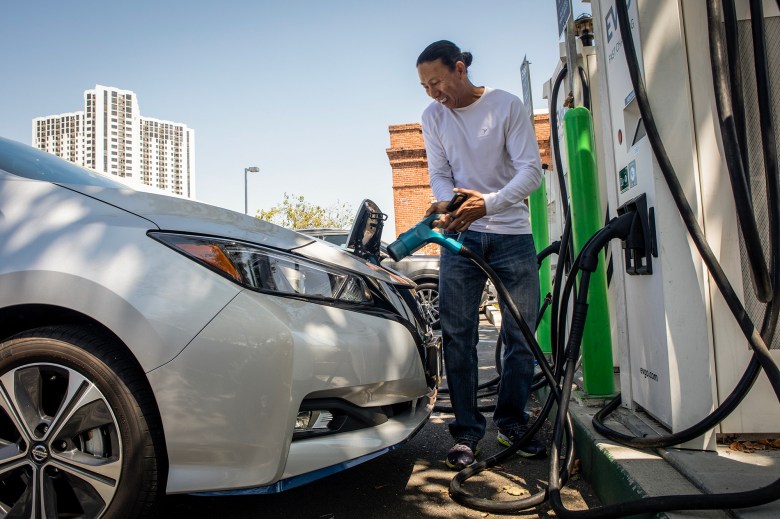 A person plugs a charging cable into an electric car at a public charging station, smiling while standing beside the vehicle as tall buildings and a brick structure rise in the background.