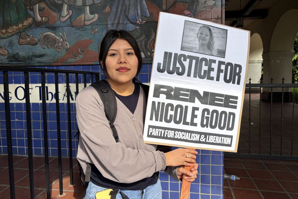 A young woman with medium light skin tone stands holding a sign that says "Justice for Renee Nicole Good."