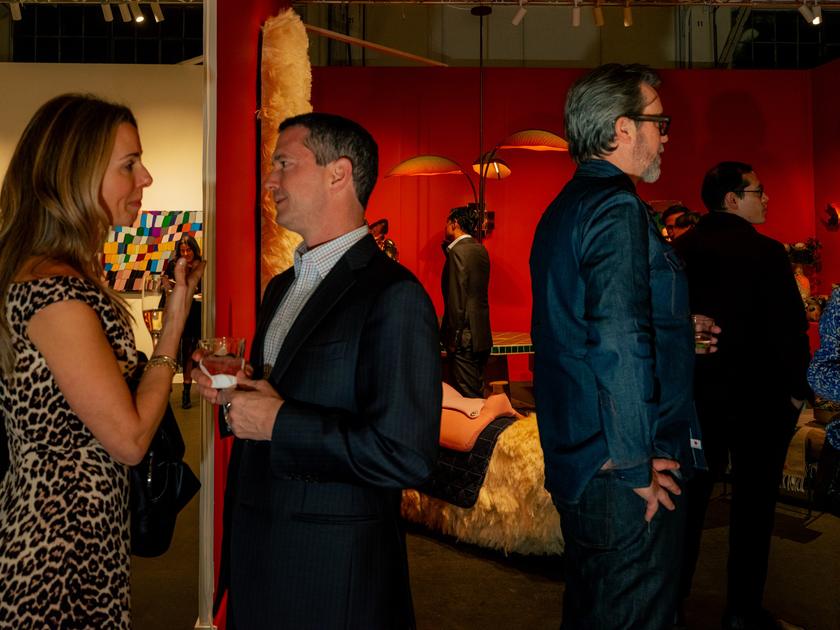Two men lean closely over a white table or pedestal, while several people in the background talk inside an art gallery.