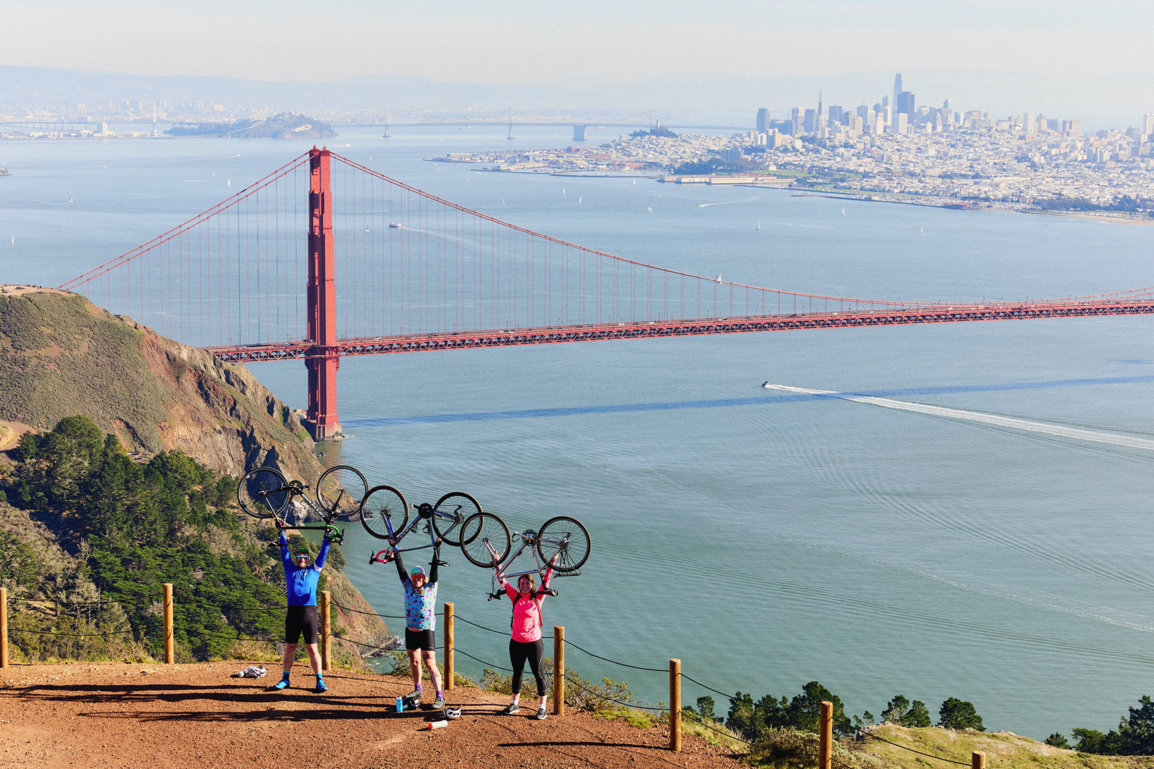 Three cyclists stand on a hill overlooking the Golden Gate Bridge, each holding their bikes above their heads with San Francisco’s skyline in the background.