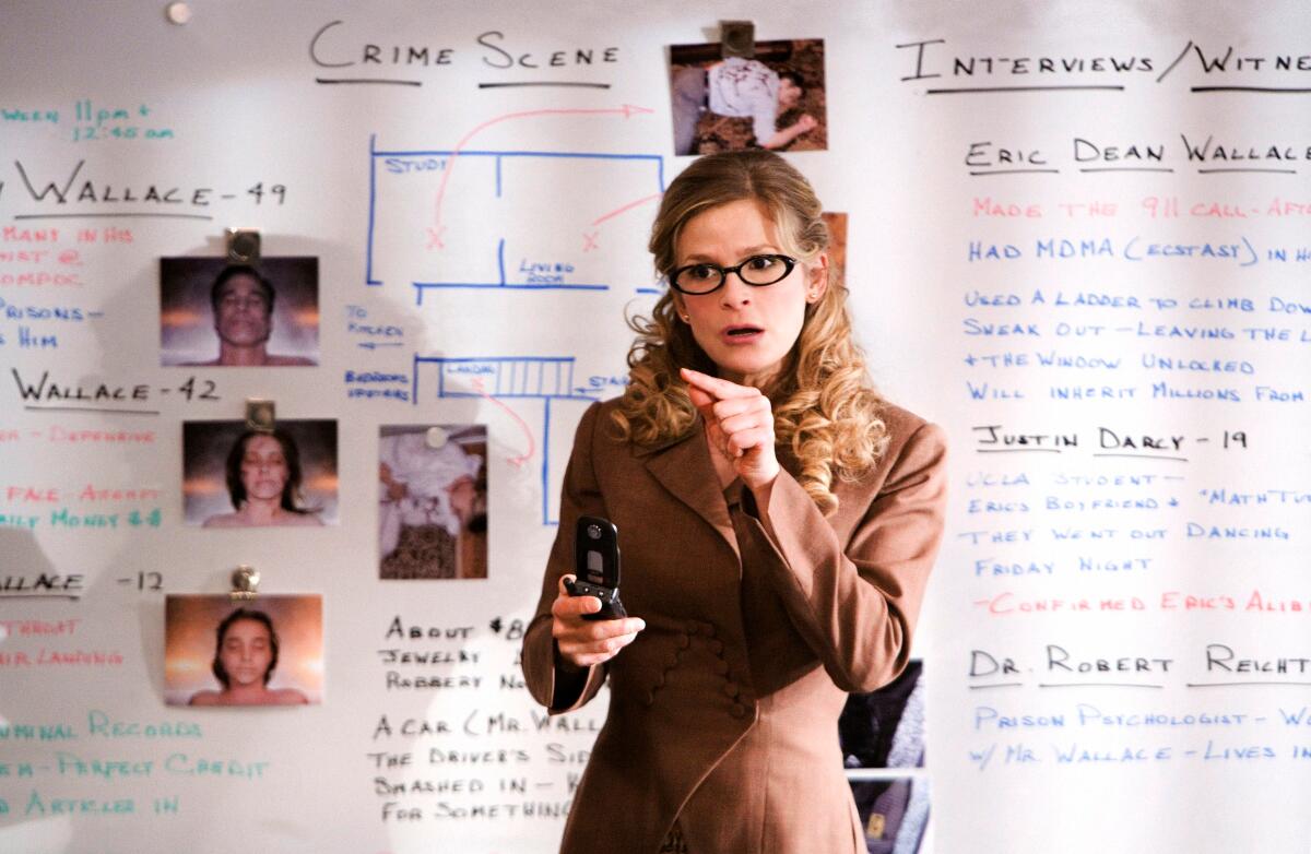 A woman wearing glasses stands in front of a white board filled with writing and taped images. 