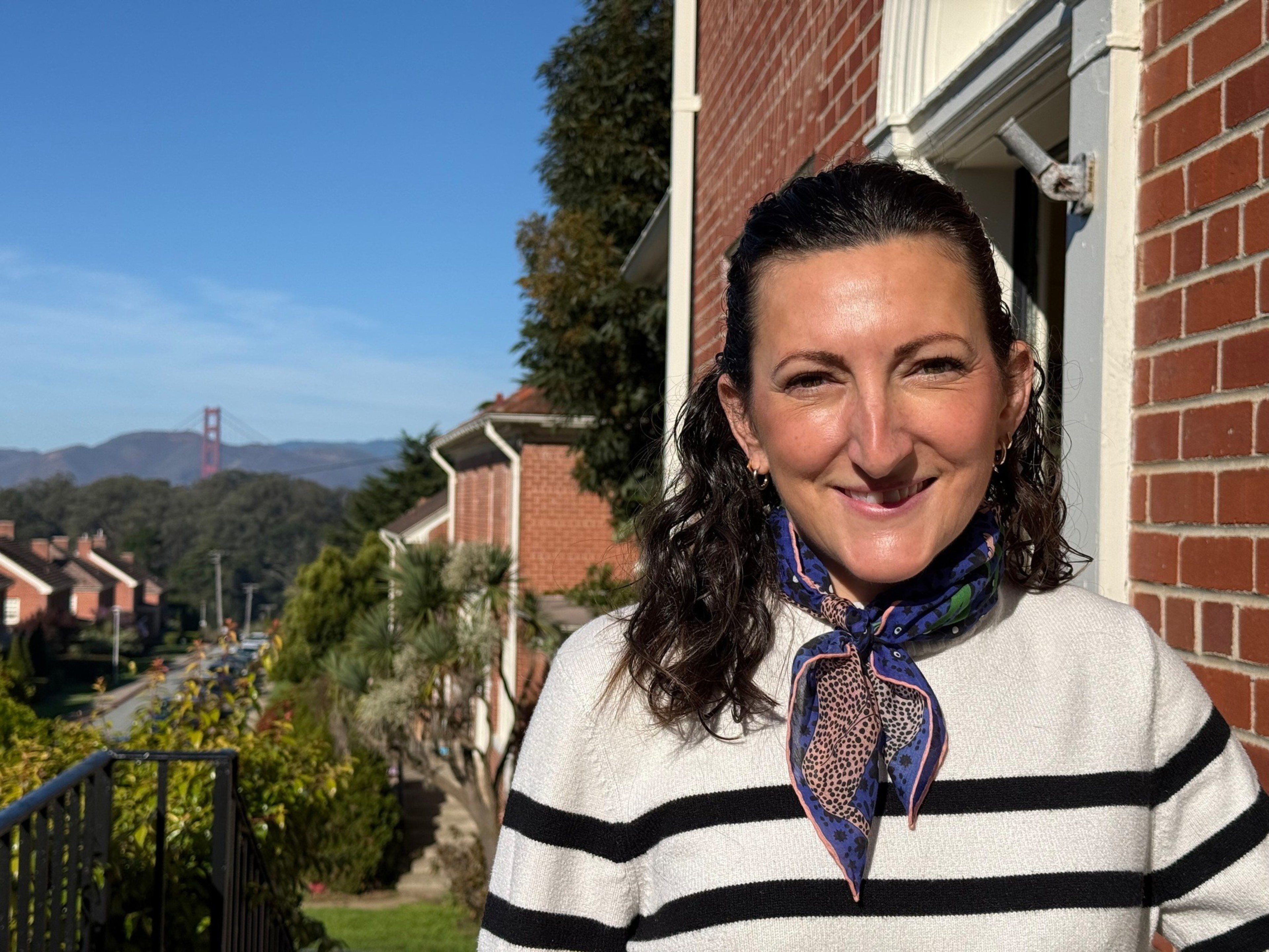 A woman with dark curly hair smiles, wearing a white sweater with black stripes and a colorful scarf, standing near a red brick building on a sunny day.
