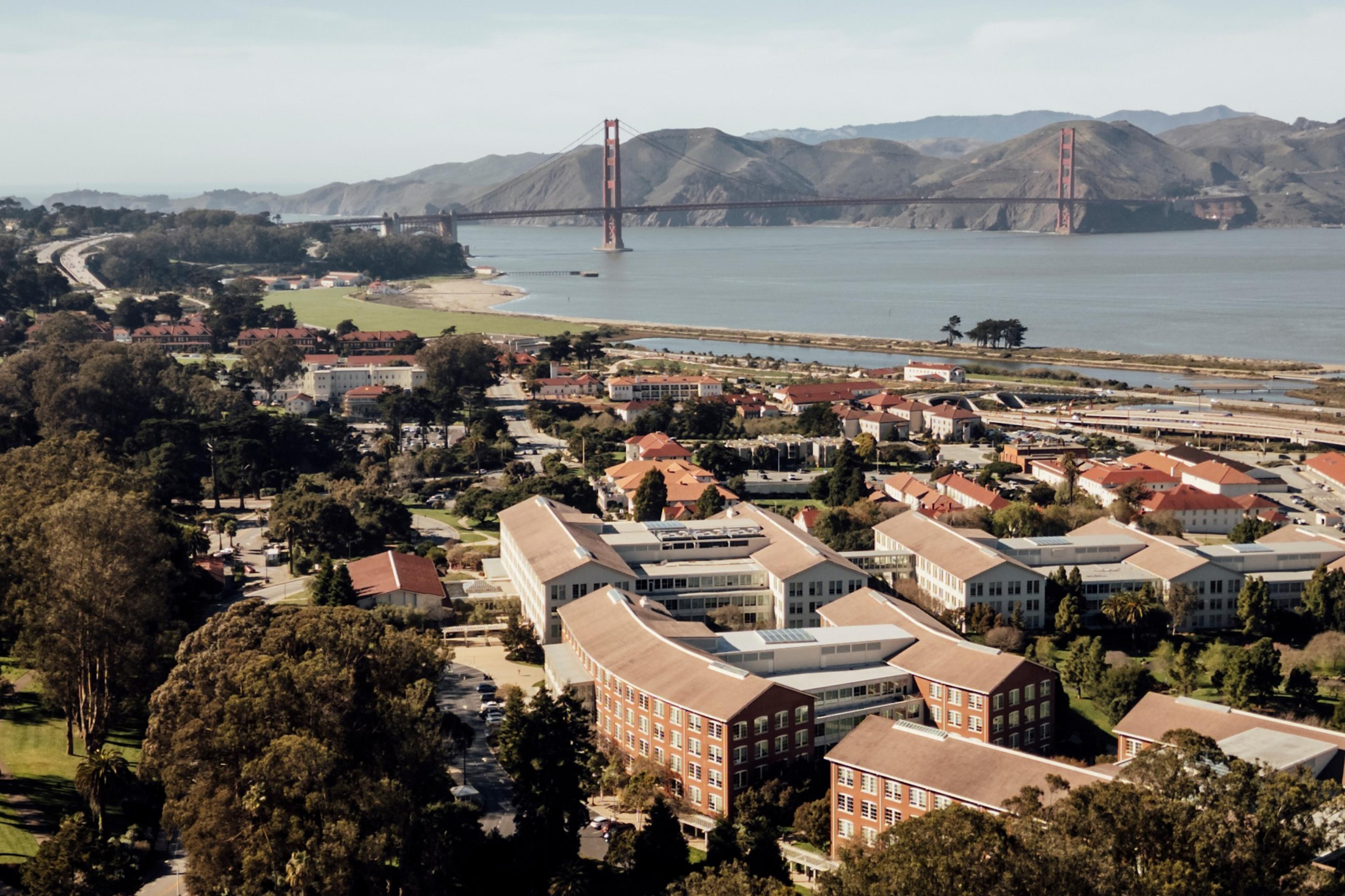 A coastal cityscape with clustered buildings and greenery in the foreground, a body of water in the middle, and the Golden Gate Bridge with hills behind it.