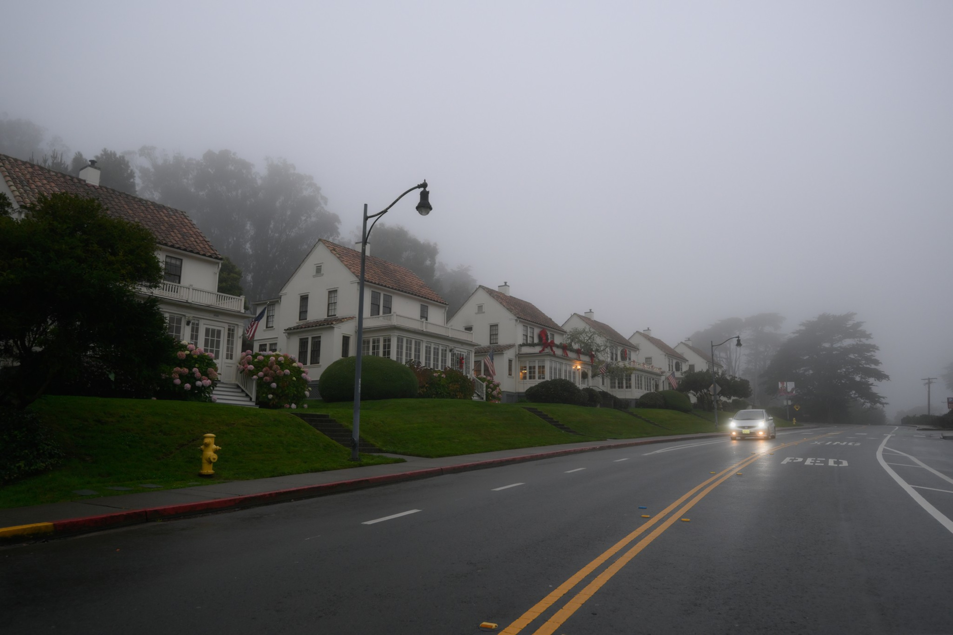 A foggy street lined with five white houses with red-tiled roofs and well-kept lawns, a car with headlights on approaches on the wet road.