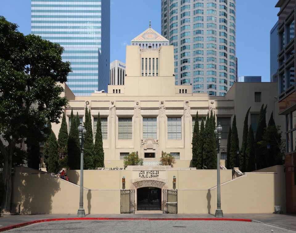 The exterior of a multi-story beige building with high-rise office towers behind it.