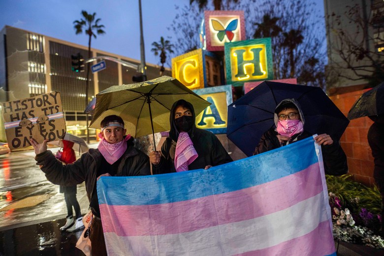 Three demonstrators stand in the rain holding umbrellas and a large transgender pride flag, with one person holding a sign reading “History has its eyes on you,” outside a brightly lit hospital sign at dusk.
