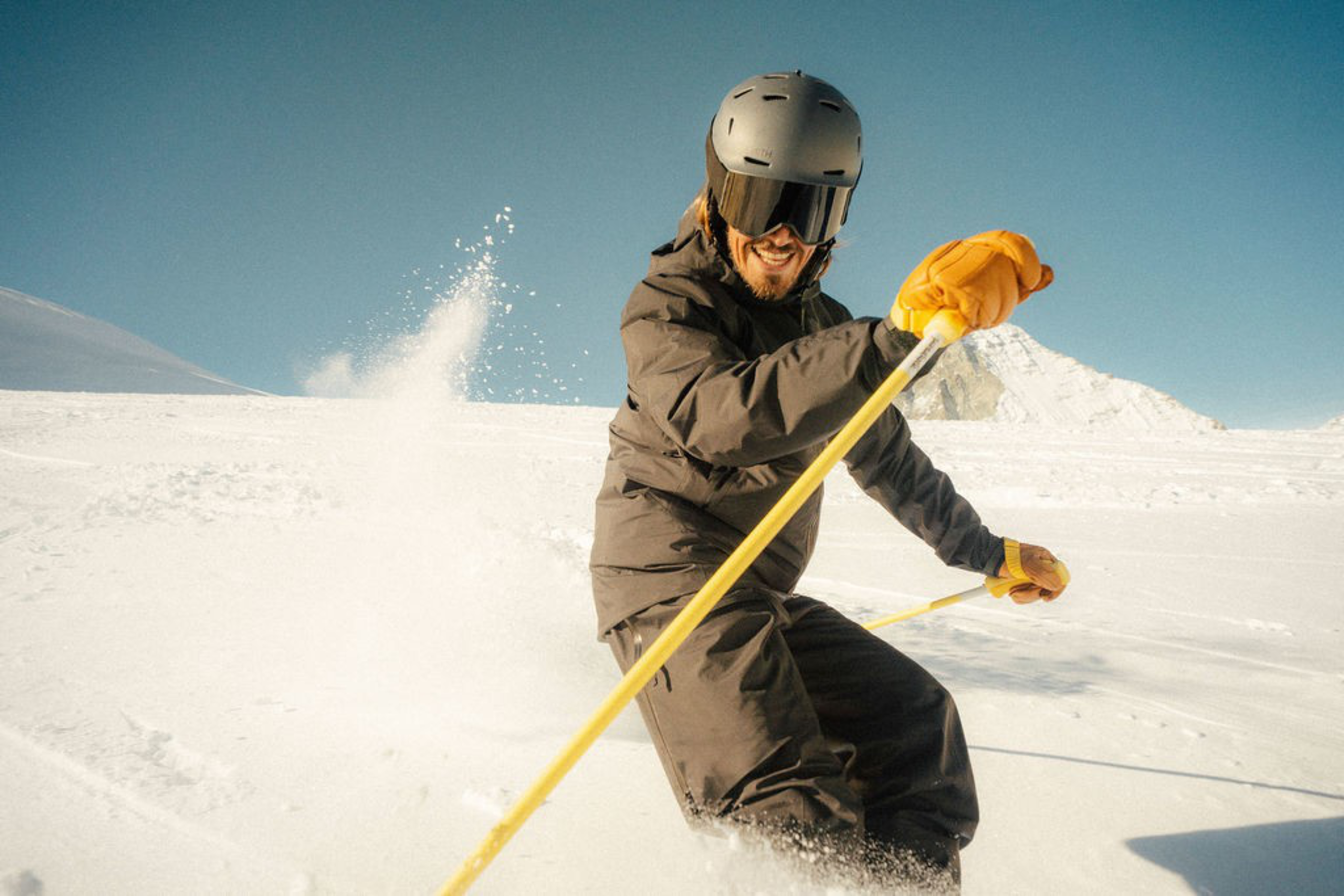 A person wearing a black helmet, goggles, and yellow gloves is skiing down a snowy slope against a clear blue sky.