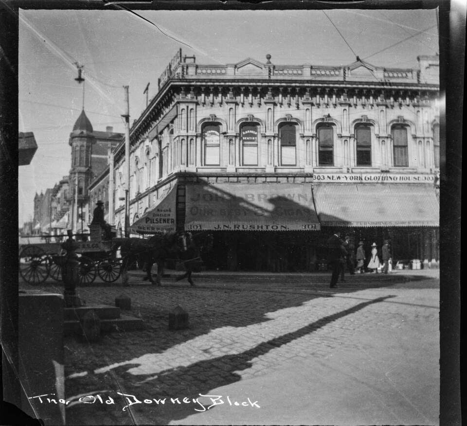 A blacka and white archival film negative of the Downey Block building on the street corner, with cobblestone streets, horse-drawn wagon, and water fountain in the foreground. The awnings and windows of the Downey Block storefronts have signs including: "Dr. Crawford, Dentist," "Dr. U.D. Reed, Dentist," "Maier & Zobelein Pilsener, Beer on Draught," "John Brown Our Best 5 Cigar ... 301 J.N. Rushton" at 301 N. Main Street, and "303 New-York Clothing House." The edge of Hazard and Harpham Patent Office can be seen attached to upper floor of building in upper left of image.