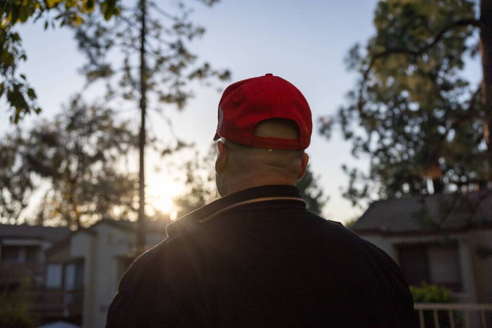 A man wearing a black jacket and red baseball cap is pictured from behind. He is facing a row of houses at sunset.