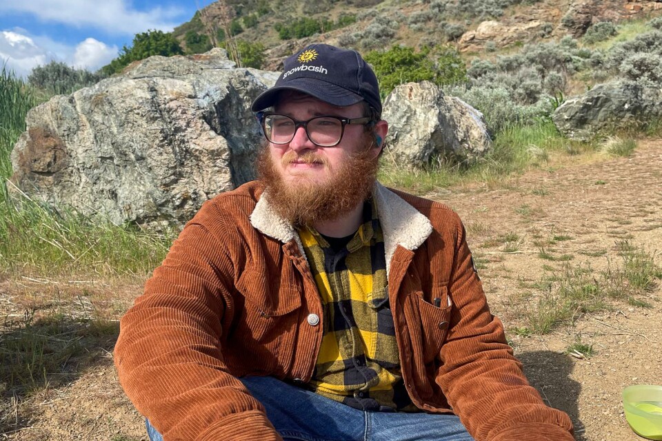 A man with a beard wearing eyeglasses a baseball cap and a brown corduroy jackets sits on the ground in front of some rocks at the base of a small hill.